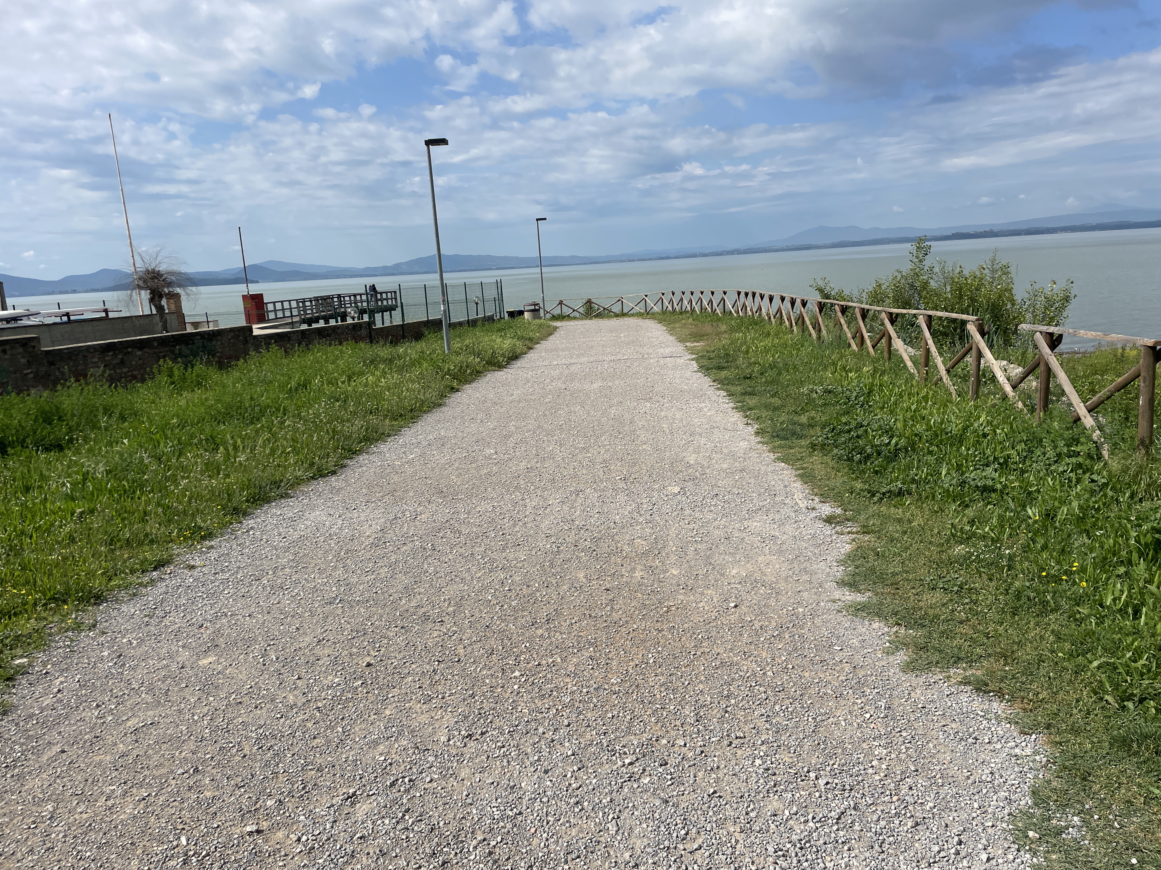 Dirt bike path at entrance to Passignano park, lined with grass and fence separating from lake.